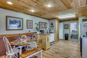Comedor con mesa de madera para cuatro, un banco y zona de cocina con refrigerador y cafetera.