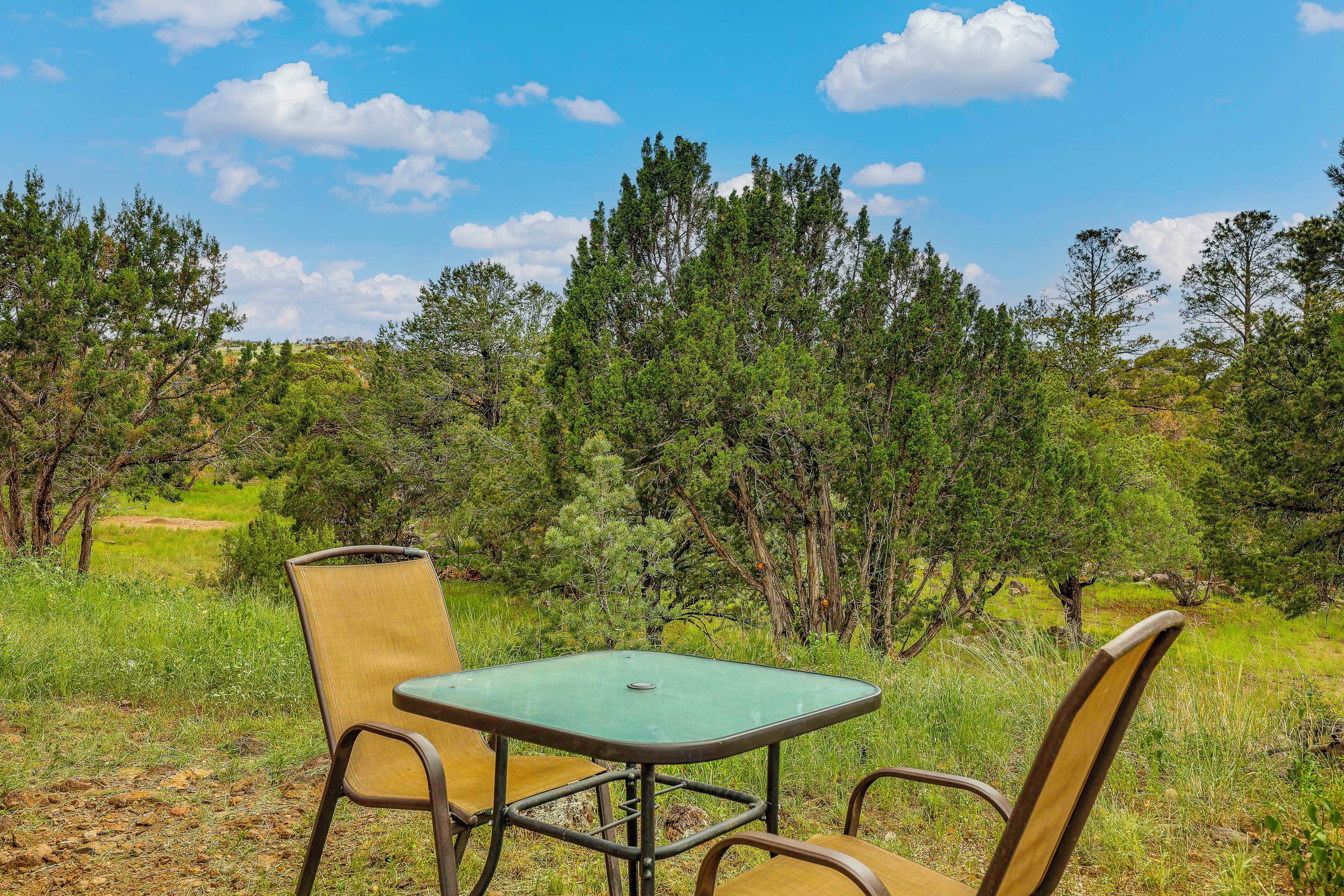 An outdoor seating area with a table and chairs, surrounded by trees and grass.