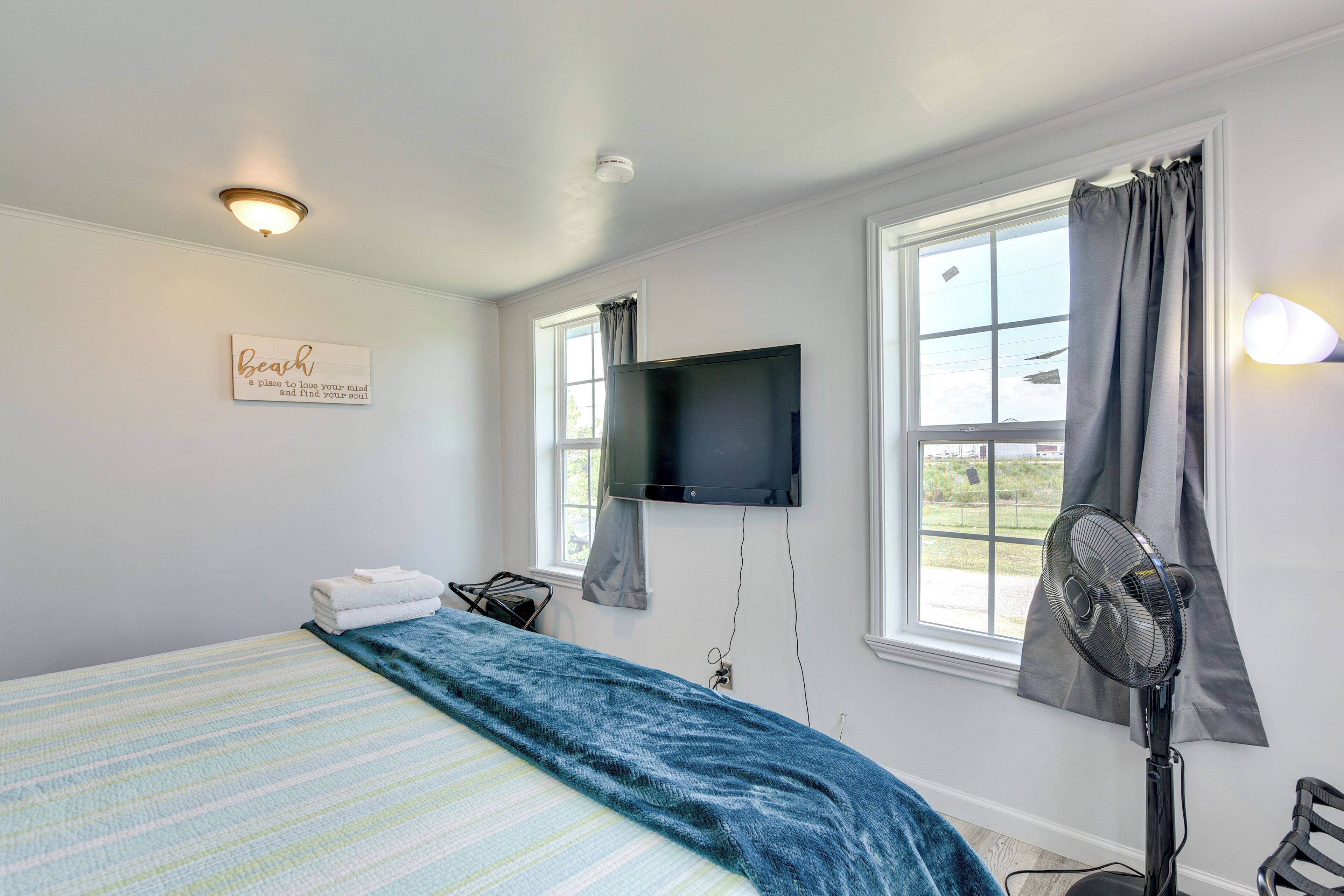 Une chambre à coucher avec un lit, un téléviseur à écran plat fixé au mur, une fenêtre avec des rideaux, un ventilateur et un panneau affiché sur le mur.