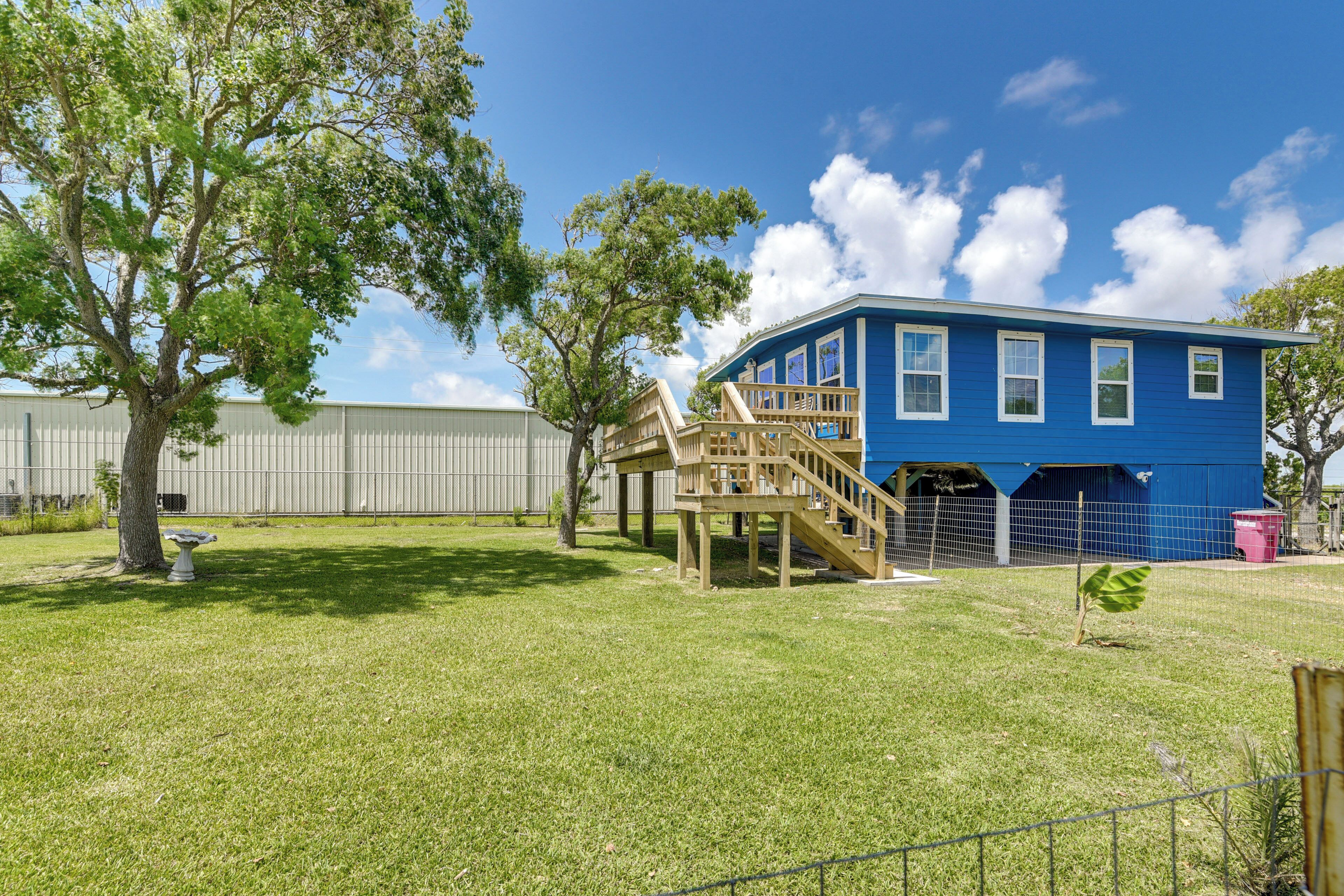 A blue house with a wooden deck and stairs, surrounded by a fenced yard with grass and trees.