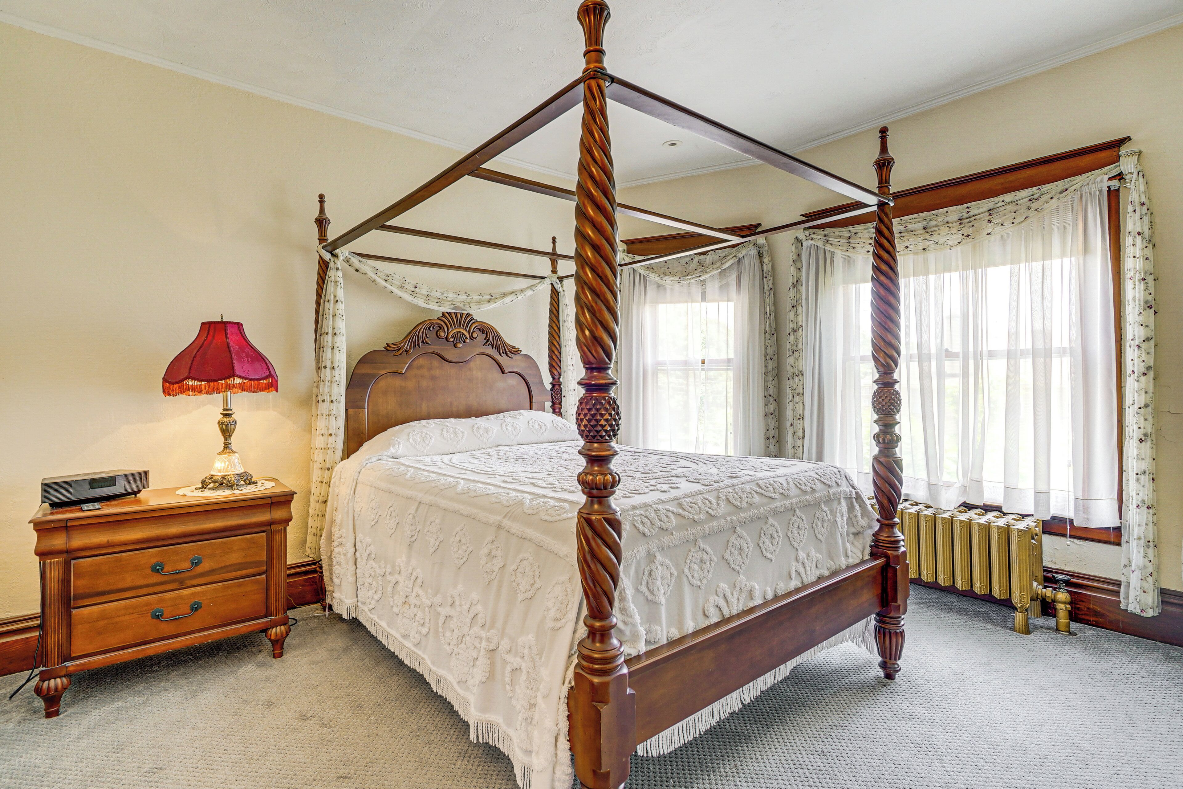 A four-poster bed with a wooden frame and a white bedspread, a wooden nightstand with a lamp, and a radiator under the window.