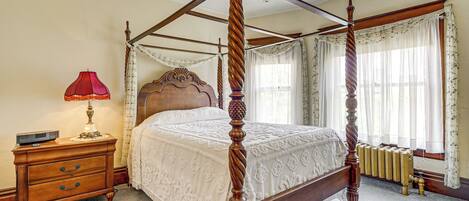A four-poster bed with a wooden frame and a white bedspread, a wooden nightstand with a lamp, and a radiator under the window.