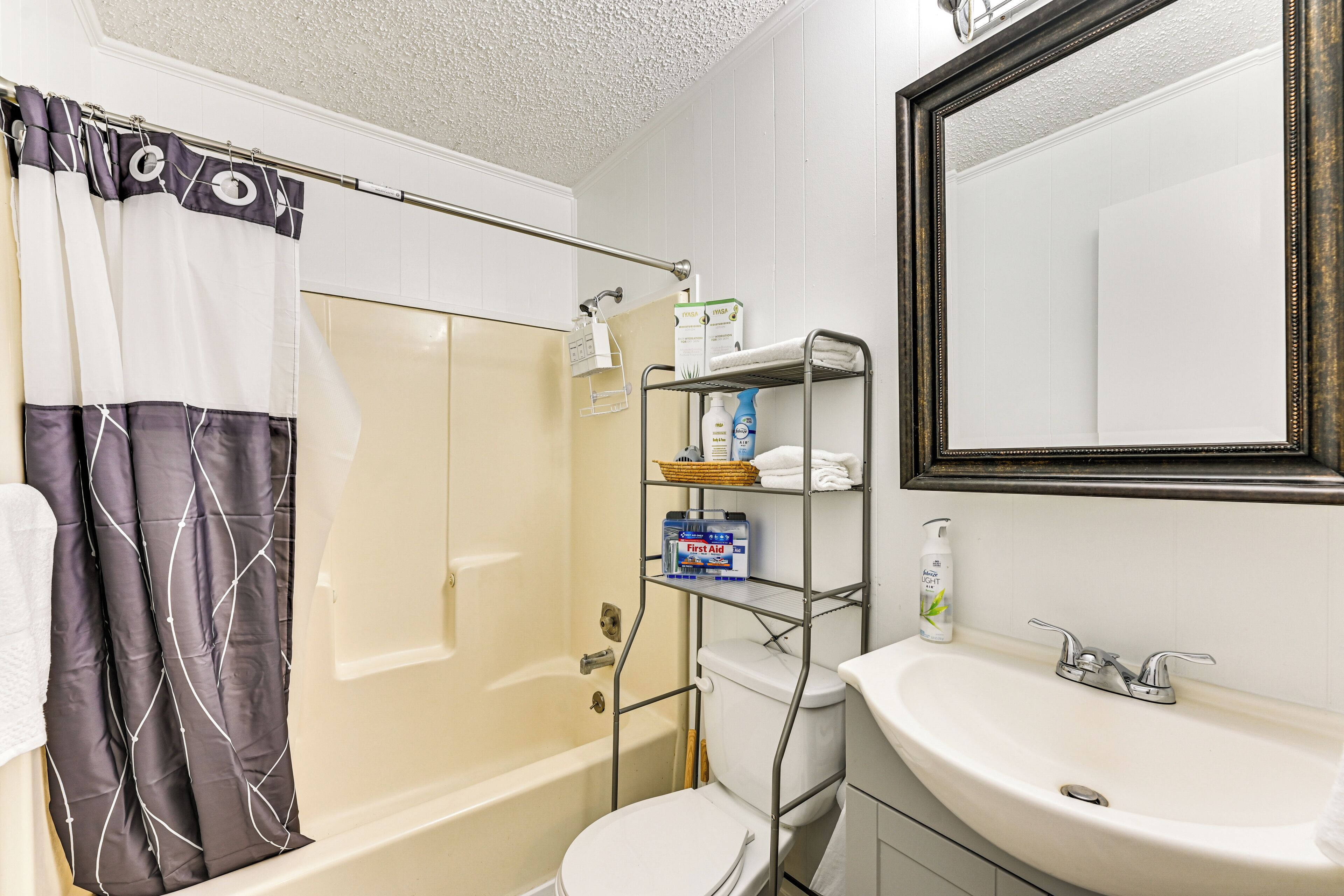 A bathroom with a bathtub, toilet, and sink. There is a shower curtain, a shelf with toiletries, and a mirror above the sink.