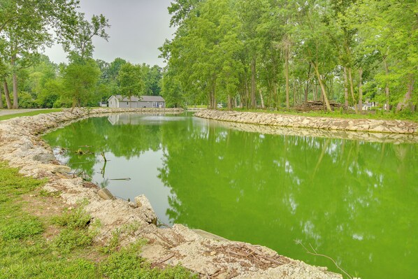 A serene pond with a stone wall and a house in the background. - Grand Lake Getaway w/ Boat Dock in St Marys! (St. Marys)