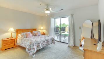 A bedroom with a bed, bedside lamps, a ceiling fan, a large window with curtains, and a wooden dresser with a mirror.