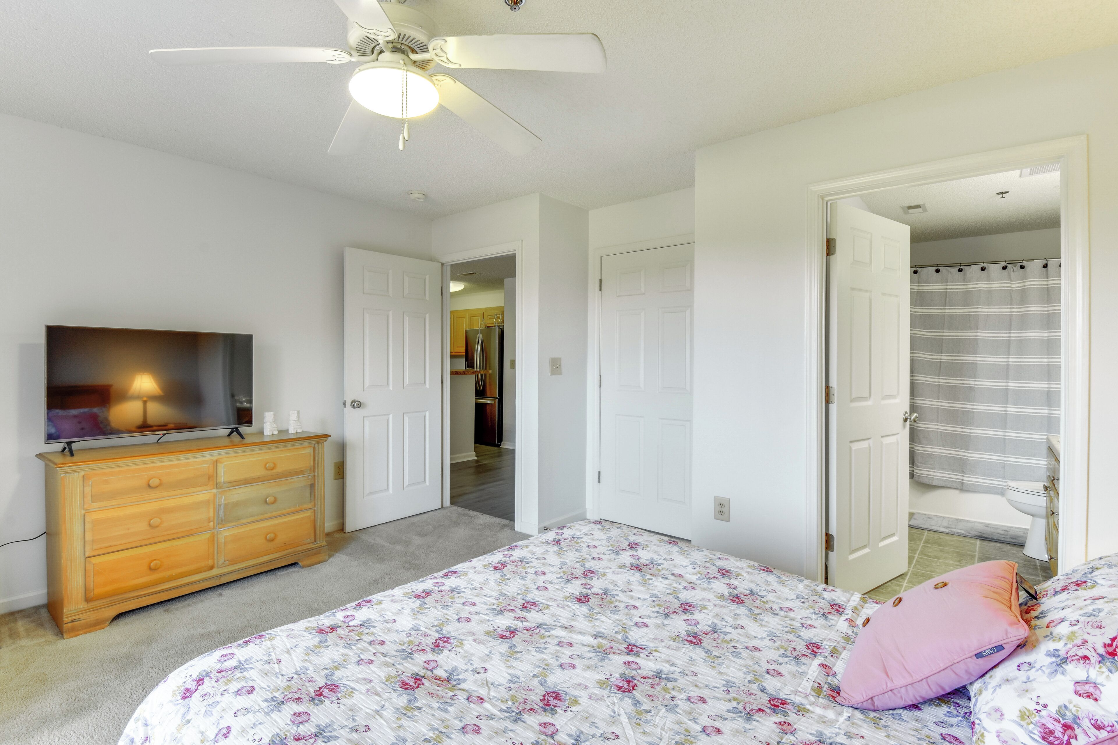 A bedroom with a bed, dresser, ceiling fan, and a door leading to a bathroom.