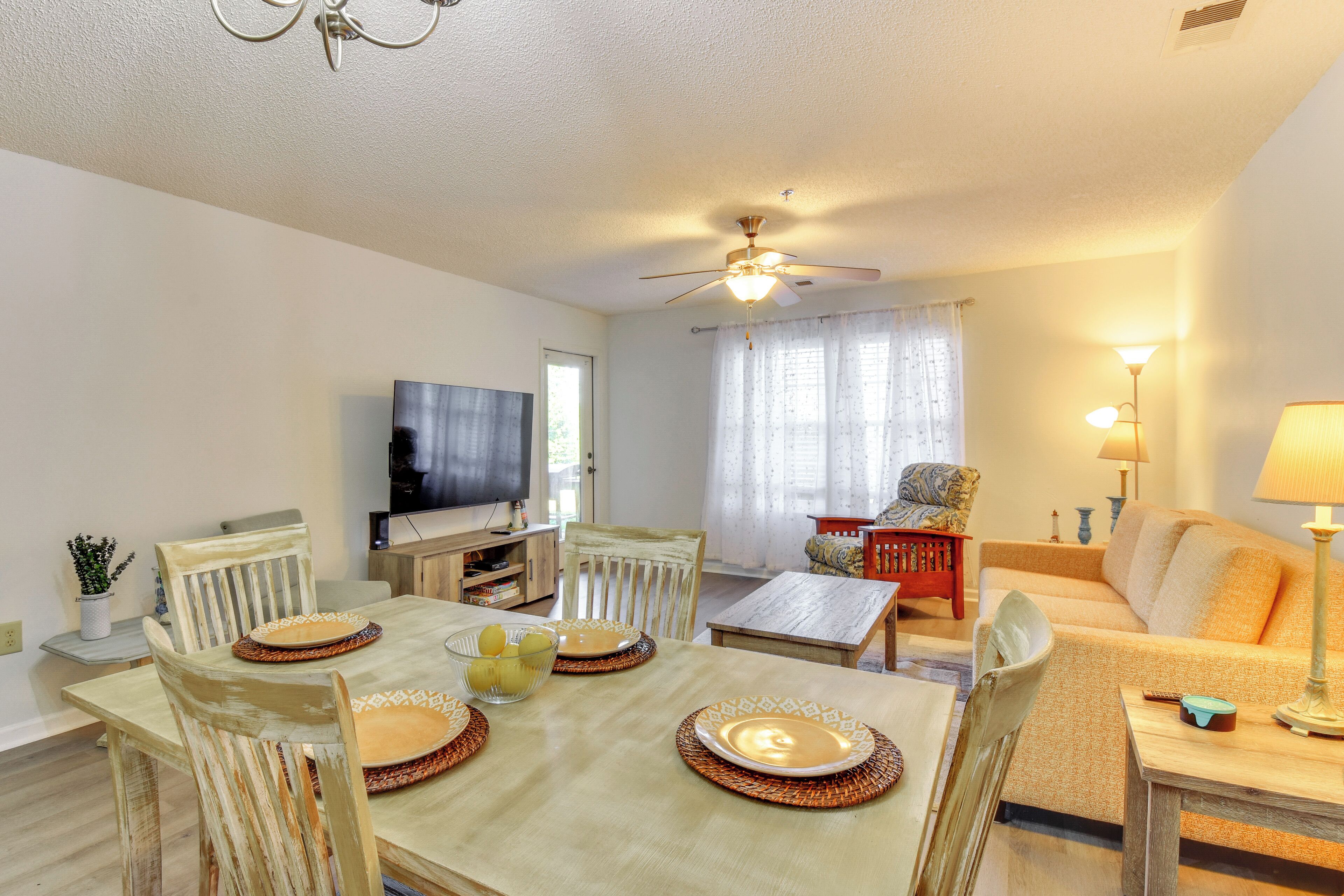 A dining area with a table set for four, surrounded by chairs. There is a television in the background, a sofa, and a lamp on a side table.