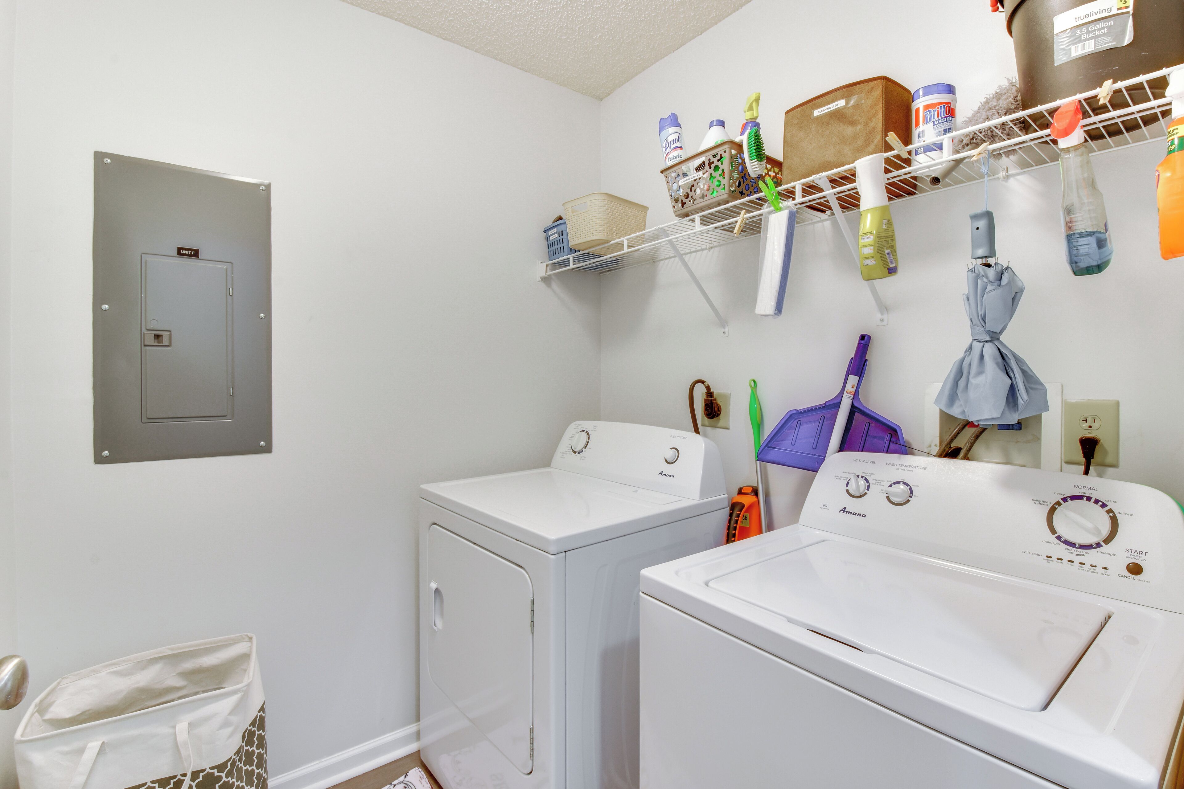 A laundry room with two white front-loading washing machines, a utility room door, and a shelf with various cleaning supplies.