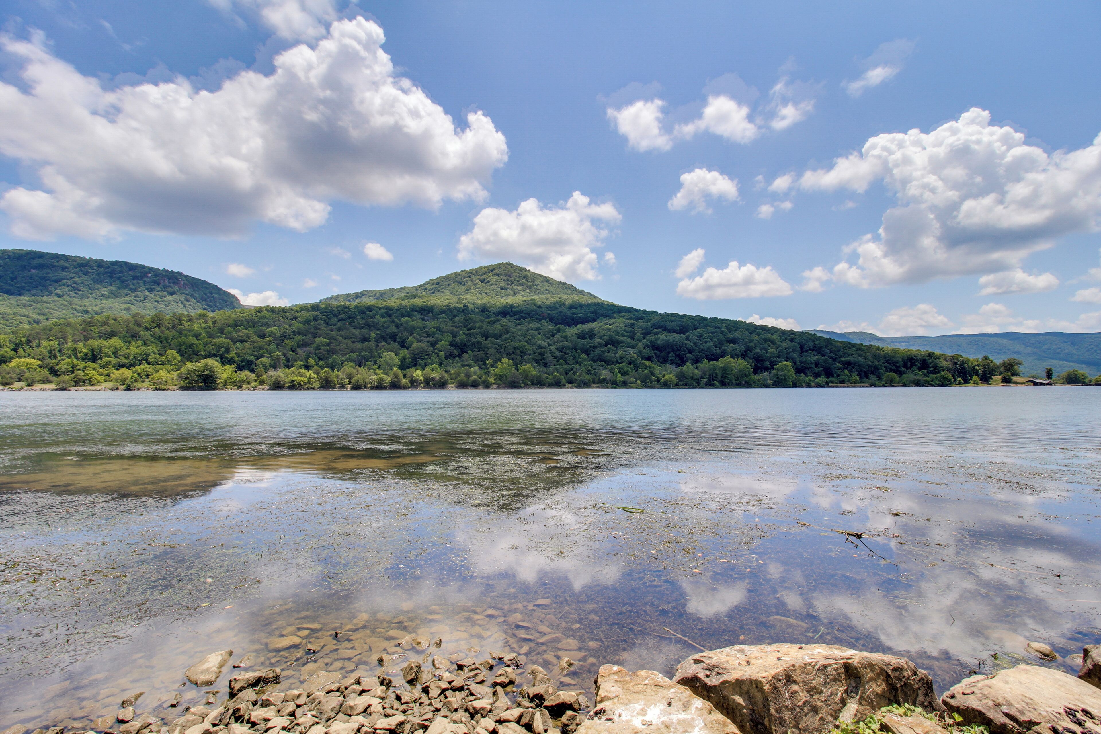 A calm lake with clear water, surrounded by lush green hills and a partly cloudy sky.