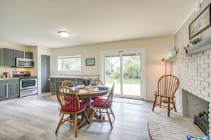 Une cuisine équipée d’appareils modernes, un coin repas avec des chaises en bois et une vue sur une maison, visible à travers une porte vitrée.
