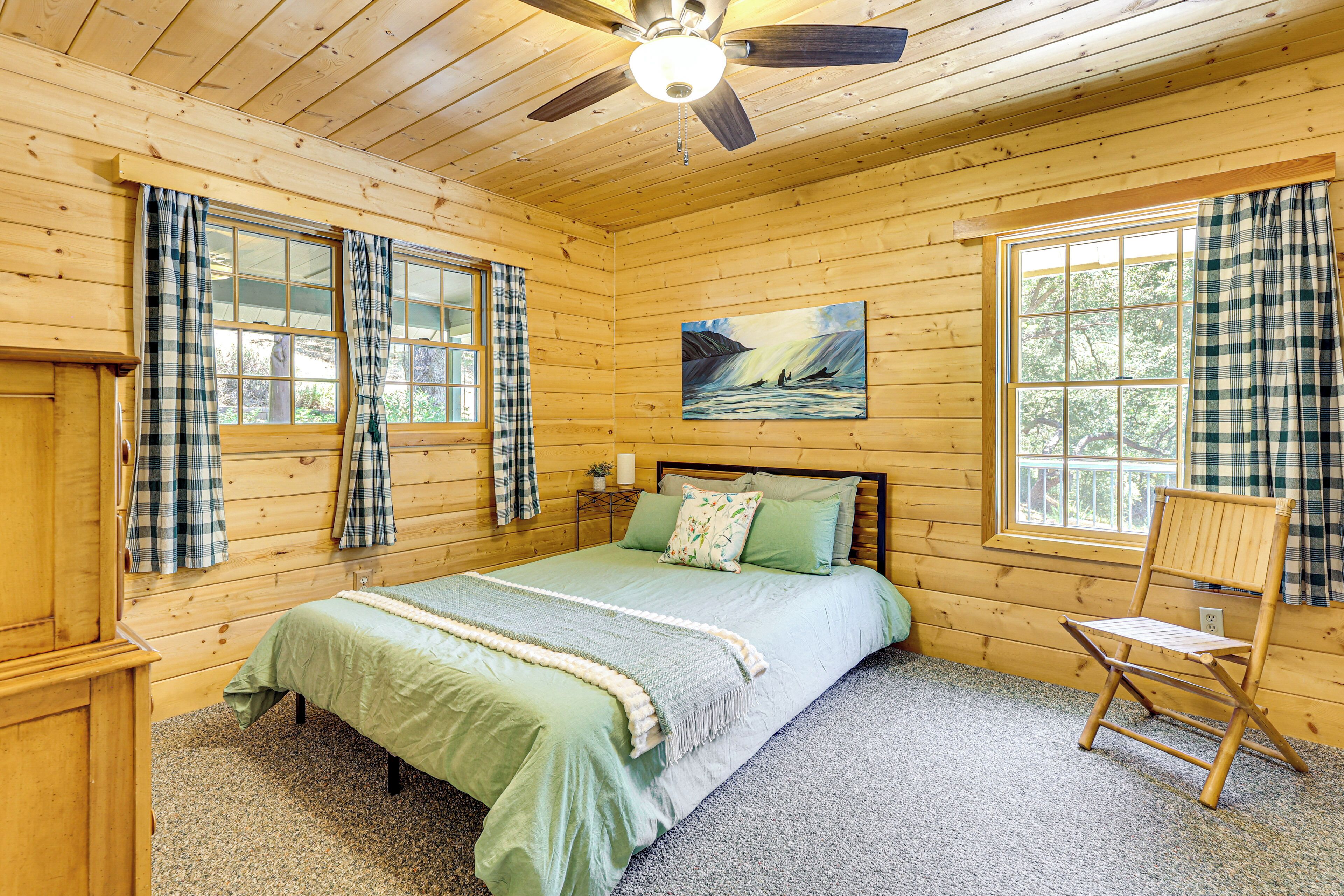 A wooden cabin bedroom with a bed, a chair, a painting, and a ceiling fan.
