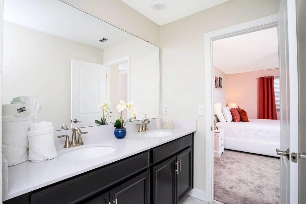 A hotel bathroom with two sinks, a large mirror, and a view into a bedroom with a bed and red curtains.
