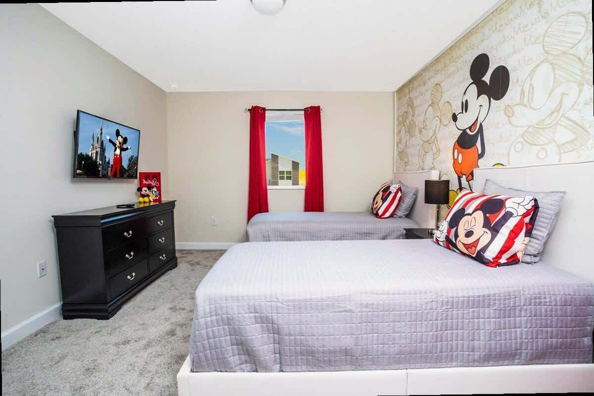 A bedroom with two beds, a dresser, a Mickey Mouse mural, and a window with red curtains.
