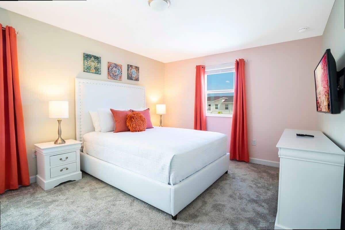 Ein Schlafzimmer mit einem weißen Bett, roten Vorhängen, einem Nachttisch mit Lampe und einem Fernseher auf einem Schrank.