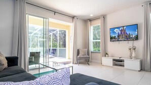 Un salon moderne avec un canapé, une table basse en verre, un téléviseur fixé au mur et un balcon offrant une vue sur la verdure.