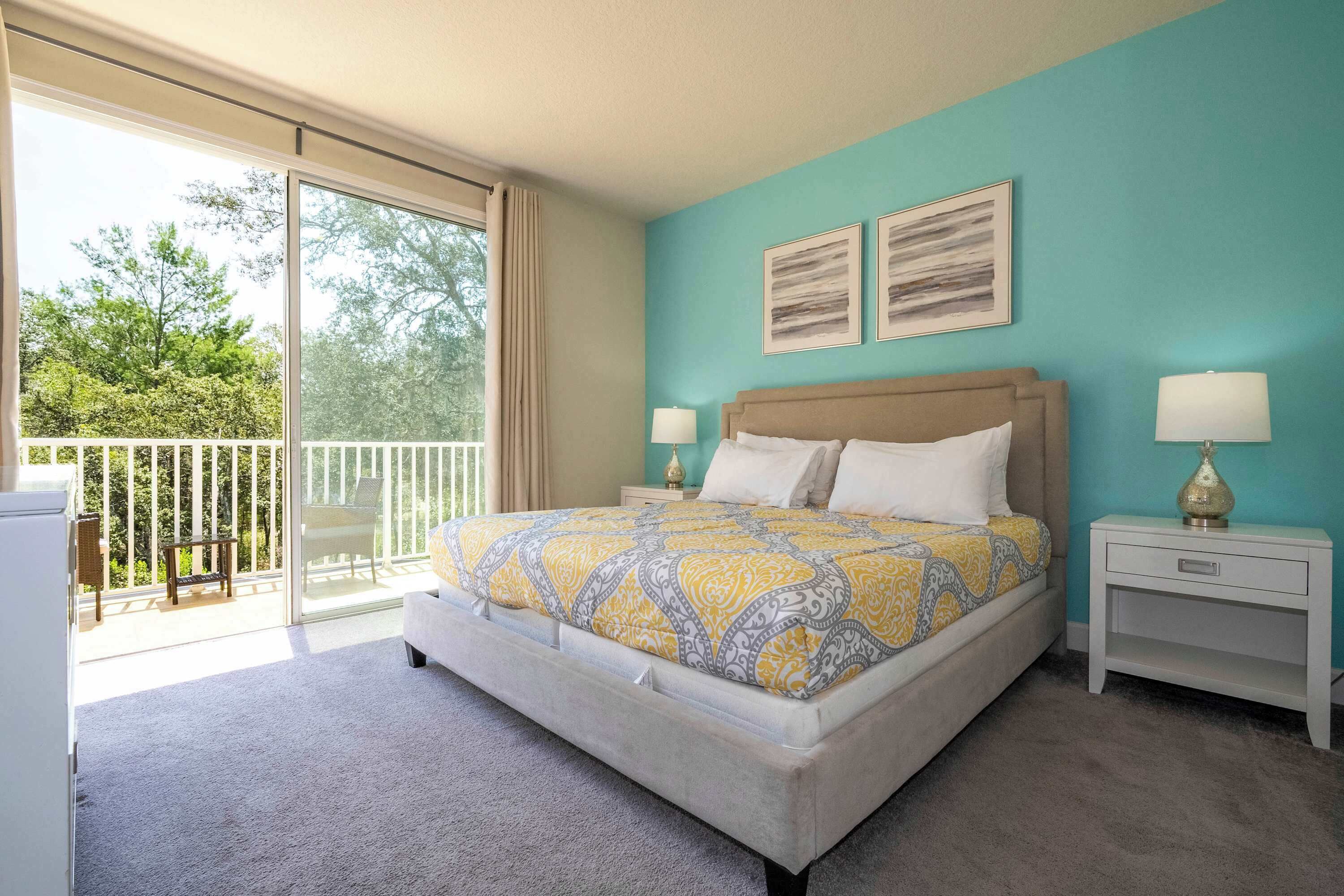 A bedroom with a large bed, two bedside tables with lamps, a view of the outdoors through a sliding glass door, and two framed pictures on the wall.