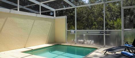 A swimming pool with a glass enclosure, surrounded by lounge chairs and a view of trees.
