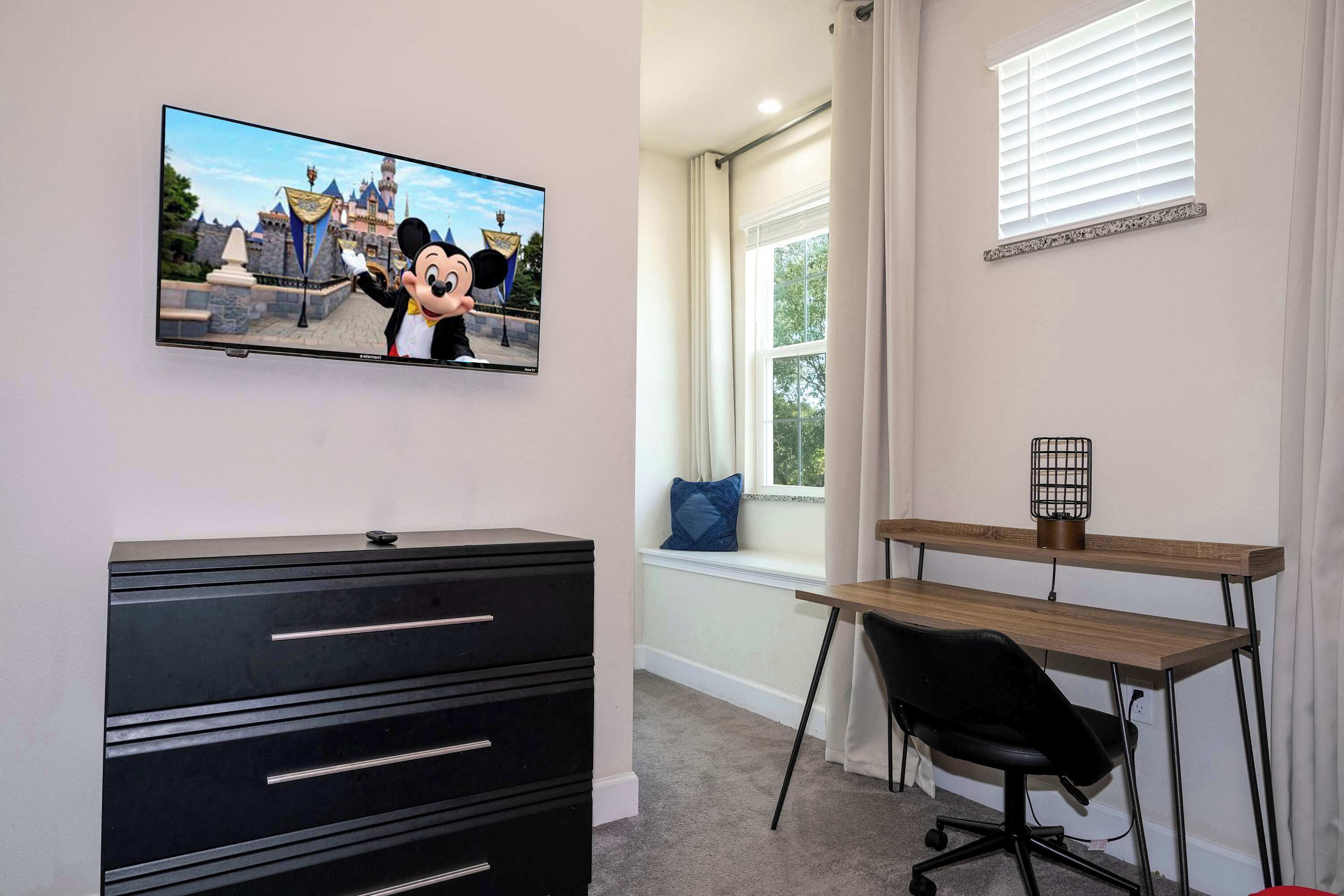 A room with a TV displaying a cartoon character, a black filing cabinet, a wooden desk, a black office chair, and a window with a view of a castle-like structure.