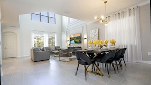 A spacious living room with a high ceiling, a dining area with a round table and black chairs, and a large window with curtains. - Themed 5Bd Family Villa w Pool Spa Near Disney (Kissimmee)