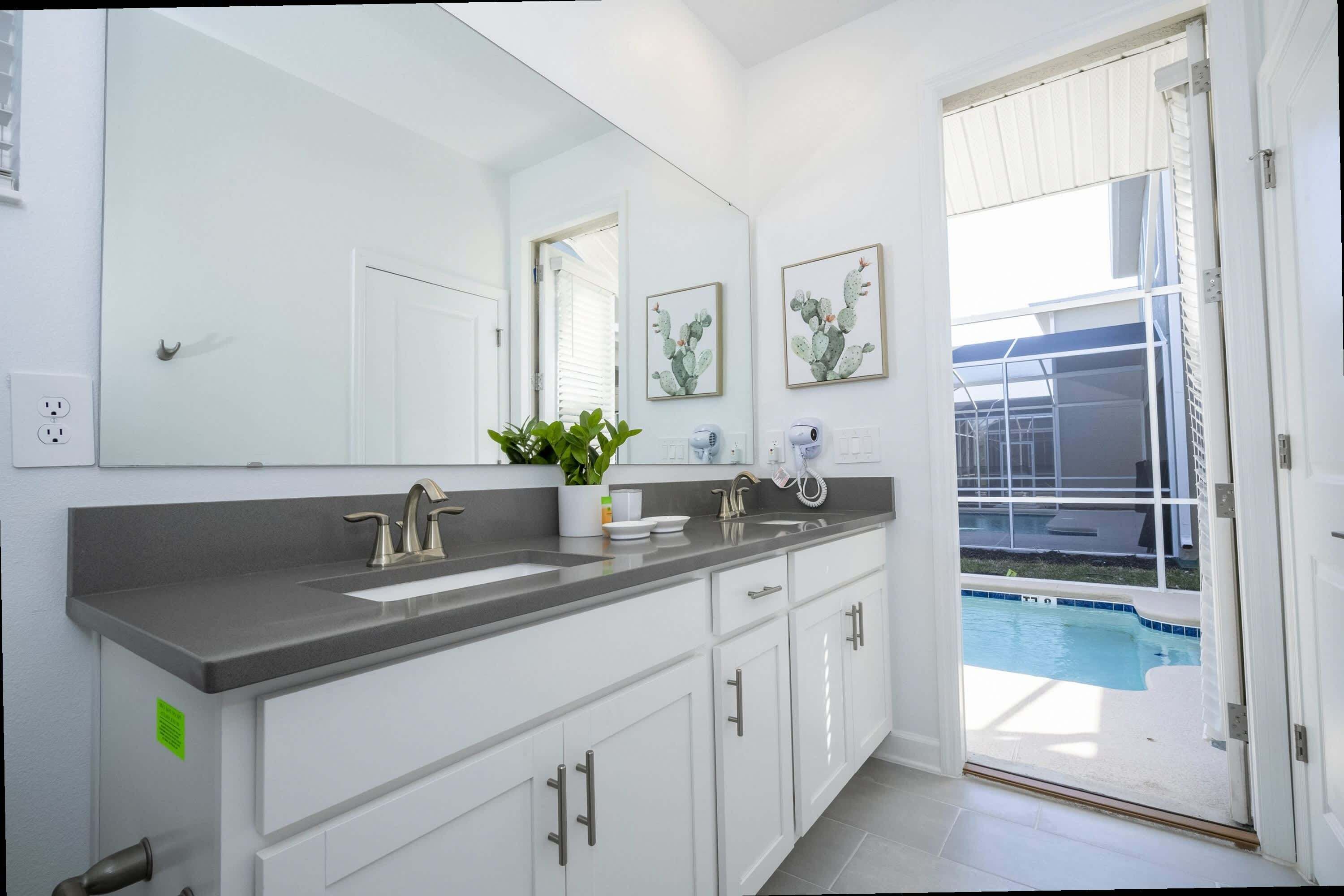A modern bathroom with a large mirror, white cabinets, a grey countertop, and a view of a swimming pool.