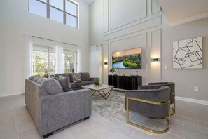 A spacious living room with a grey sofa set, a coffee table, and a TV mounted on the wall. - Themed 5Bd Family Villa w Pool Spa Near Disney (Kissimmee)