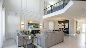 A spacious living room with a grey sofa set, a glass coffee table, and a painting of a landscape. - Themed 5Bd Family Villa w Pool Spa Near Disney (Kissimmee)