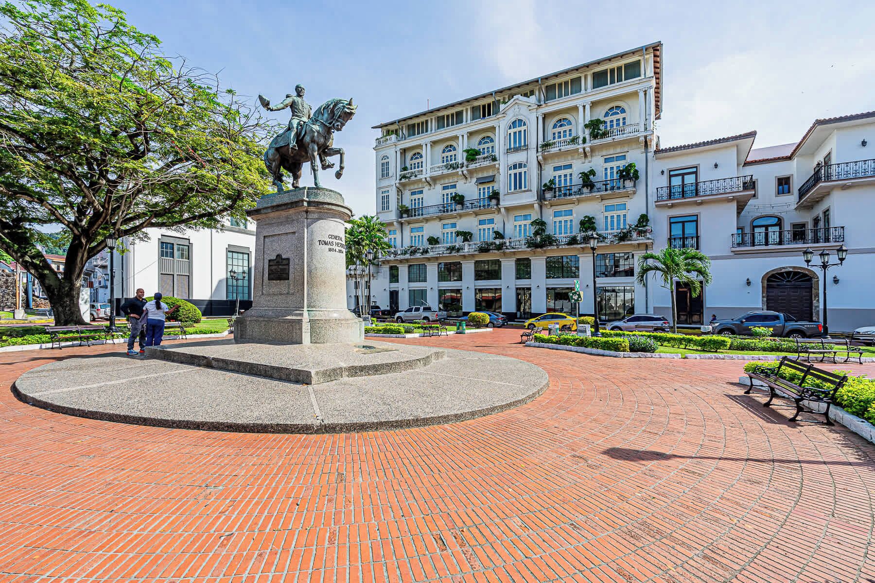 A plaza with a statue, a building with balconies, and a paved walkway.