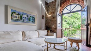 A living room with a beige sofa, a round glass coffee table, and a wicker side table. A painting of houses is on the wall, and a stone wall is visible. - w Grandiose 1BR w Balcony in San Felipe (Panama City)