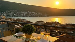 A table set for two with glasses of white wine, a bowl, and a potted plant on a balcony overlooking a body of water and a town. - Panoramic Seaview Blue Nest - Stylish Getaway (Amfilochia)