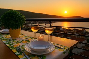 Mesa dispuesta con dos copas de vino, una vela y un bol en un balcón con vista a la puesta de sol sobre el agua.