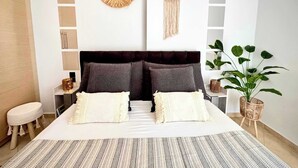 A modern bedroom with a large bed, two pillows, a headboard, a bedside table, a small stool, a wall-mounted macrame piece, a potted plant, and a patterned rug. - Panoramic Seaview Blue Nest - Stylish Getaway (Amfilochia)
