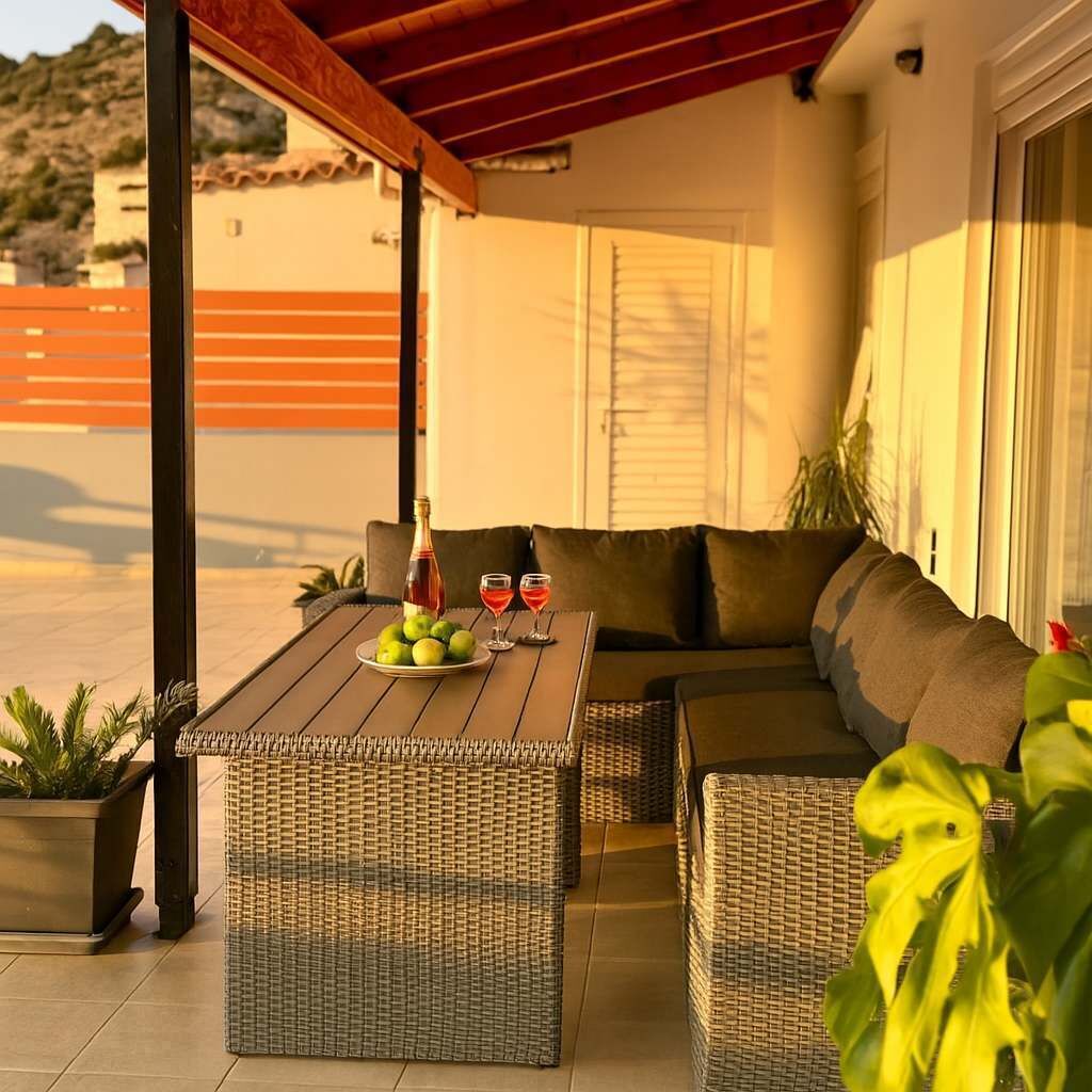 A covered outdoor seating area with wicker furniture, a table with a bottle and glasses, and a potted plant.