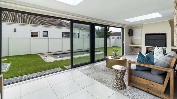 A spacious living room with a large sliding glass door leading to a backyard, a comfortable seating area with a sofa and coffee table, and a flat-screen TV mounted on the wall. - Spacious Sunningdale Retreat With Poolside Charm (Cape Town)