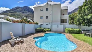 A swimming pool with a brick border, a white building with balconies, and a mountain in the background. - 2 Bedroom Ground Floor Apartment Princess Beach Club Houtbay (Cape Town)