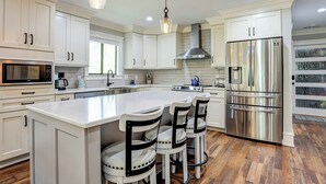 A modern kitchen with white cabinetry, stainless steel appliances, a central island with bar stools, and a hanging light fixture. - Sunset Rentals | 8 Governors Lane (Hilton Head Island)