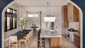 A modern kitchen with a central island, wooden cabinets, and a dining area with chairs and a table. - Modern NOLA Homes | Heated Pools & Spas (New Orleans)