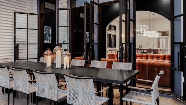 A dining area with a dark table and wicker chairs, large windows, and a view into a living room with orange furniture. - Modern NOLA Homes | Heated Pools & Spas (New Orleans)