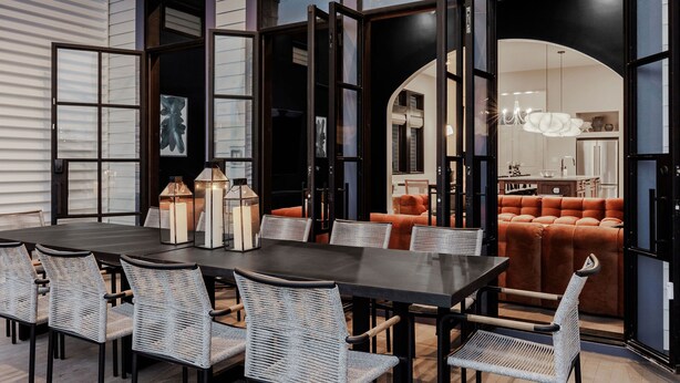 Een eethoek met een donkere tafel en rieten stoelen, grote ramen en uitzicht op een woonkamer met oranje meubels.