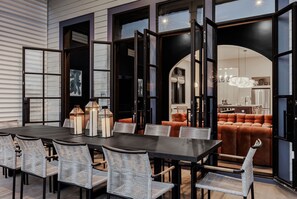 A dining area with a dark table and wicker chairs, large windows, and a view into a living room with orange furniture. - Modern NOLA Homes | Heated Pools & Spas (New Orleans)
