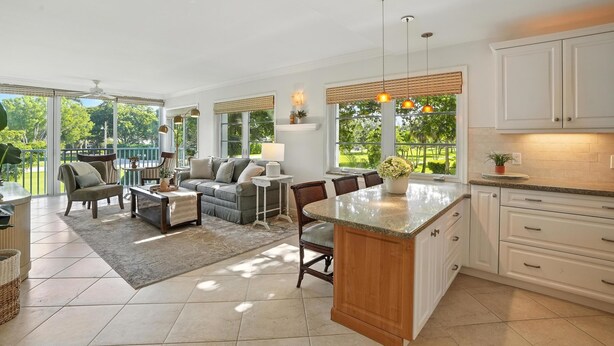 A spacious living area with a large sofa, chairs, and a coffee table. There is a kitchen area with white cabinets and a large island. The room has large windows with blinds, allowing natural light to enter. - Olde Naples Condo – Walk to Beach & Dining (Naples)