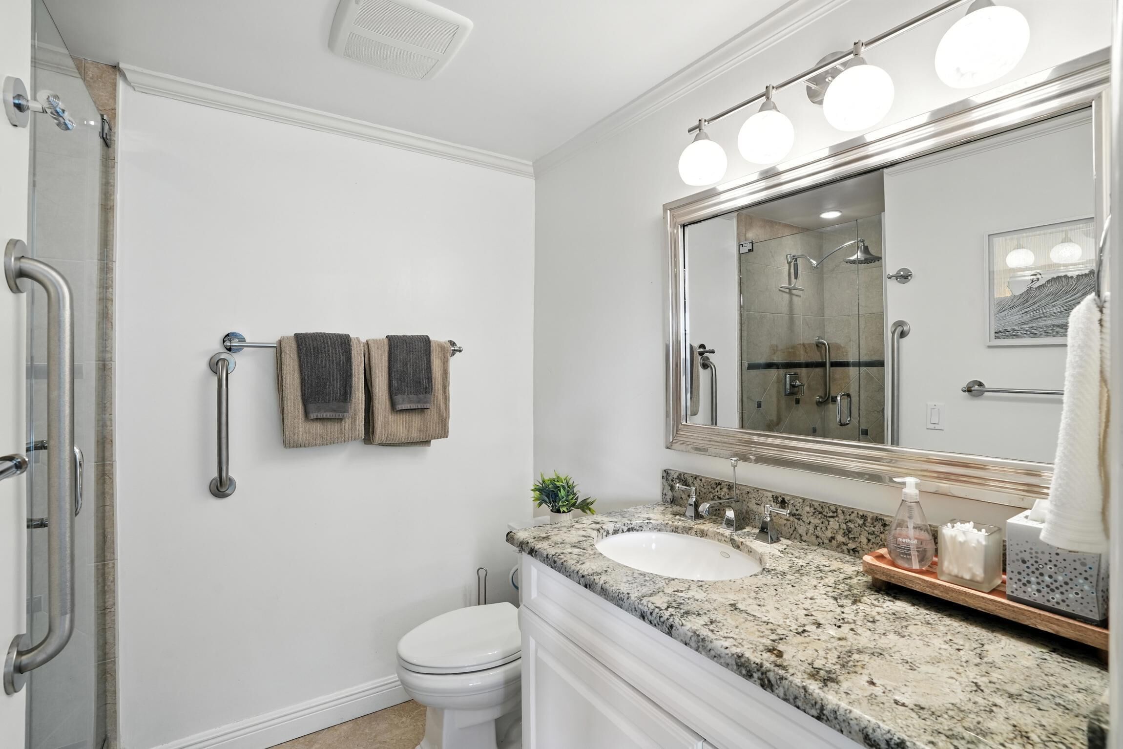 A bathroom with a white toilet, a sink with a granite countertop, a large mirror with lights, and a shower area.