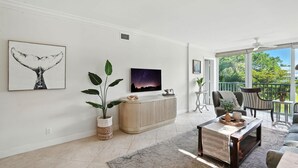 A modern living room with a large window, a ceiling fan, a sofa, a coffee table, and a TV. - Olde Naples Condo – Walk to Beach & Dining (Naples)