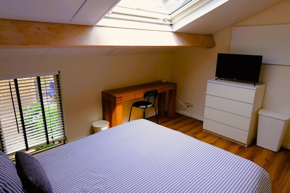 Un dormitorio con cama, escritorio y silla, televisor en una cómoda, y tragaluz.