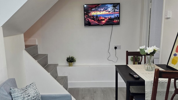 Una sala de estar moderna con un sofá gris, una mesa de comedor con sillas, un televisor montado en la pared y una escalera que sube a otro nivel.