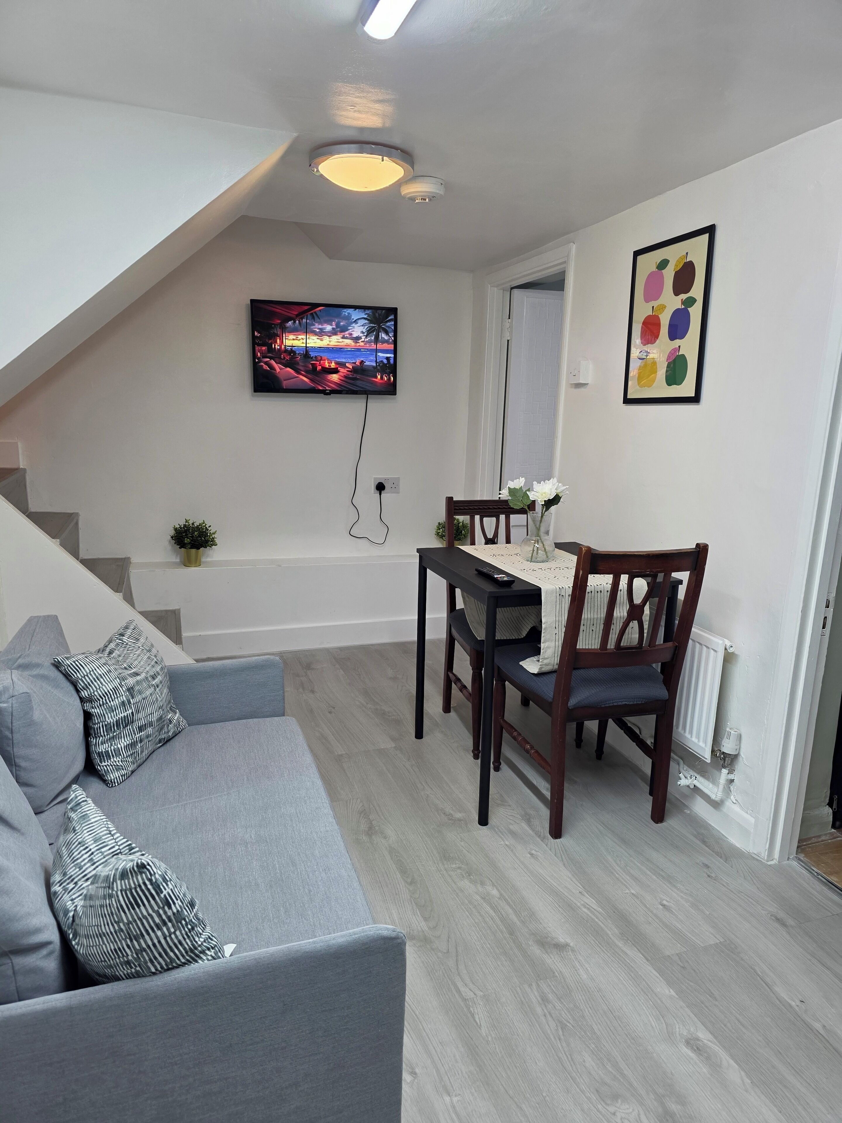 A modern living room with a grey sofa, a dining table with chairs, a TV mounted on the wall, and a staircase leading to an upper level.