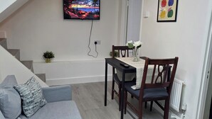 Una sala moderna con un sofá gris, una mesa de comedor con sillas, un televisor montado en la pared y una escalera que sube a un nivel superior.