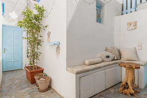 A narrow alleyway with a blue door, a potted plant, a bench with cushions, and a wooden table.