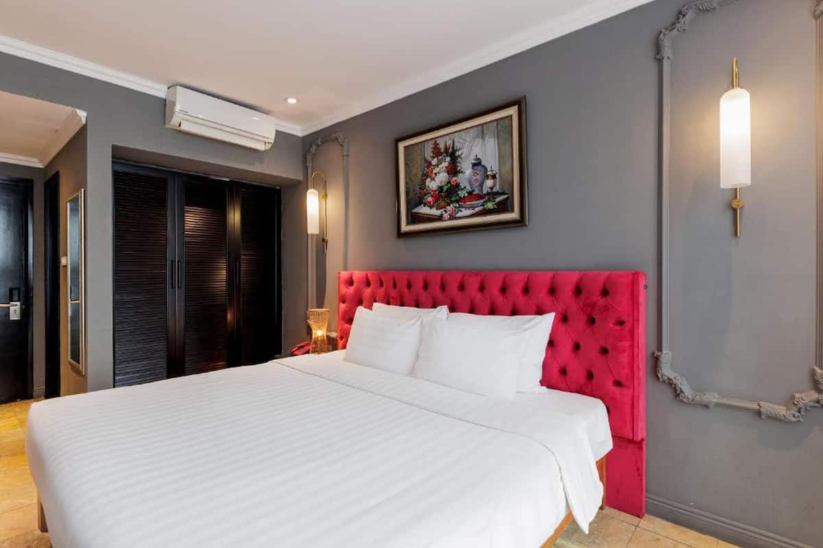 Une chambre d’hôtel avec un grand lit, une tête de lit rouge à motifs capitonnés, un tableau au mur et un luminaire fixé au mur.
