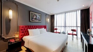 Ein Hotelzimmer mit einem großen Bett, einem roten Kopfteil, einem Gemälde über dem Bett, einem Schreibtisch mit Stuhl und einem großen Fenster mit Vorhängen.