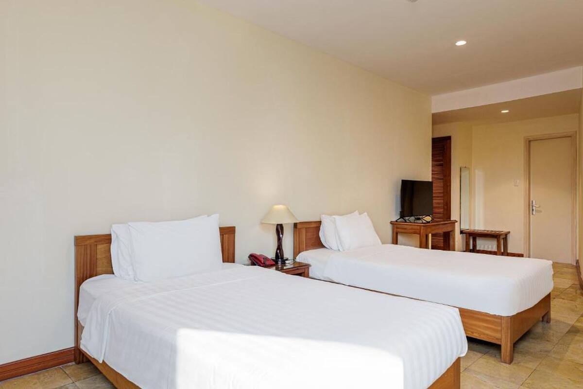 Une chambre d’hôtel avec deux lits simples, une table de chevet avec une lampe, une télévision et une petite table d’appoint en bois.
